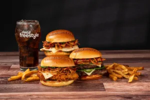 Three BurgerFi burgers with crispy toppings, served alongside golden fries and a refreshing Coca-Cola Freestyle drink on a wooden table.