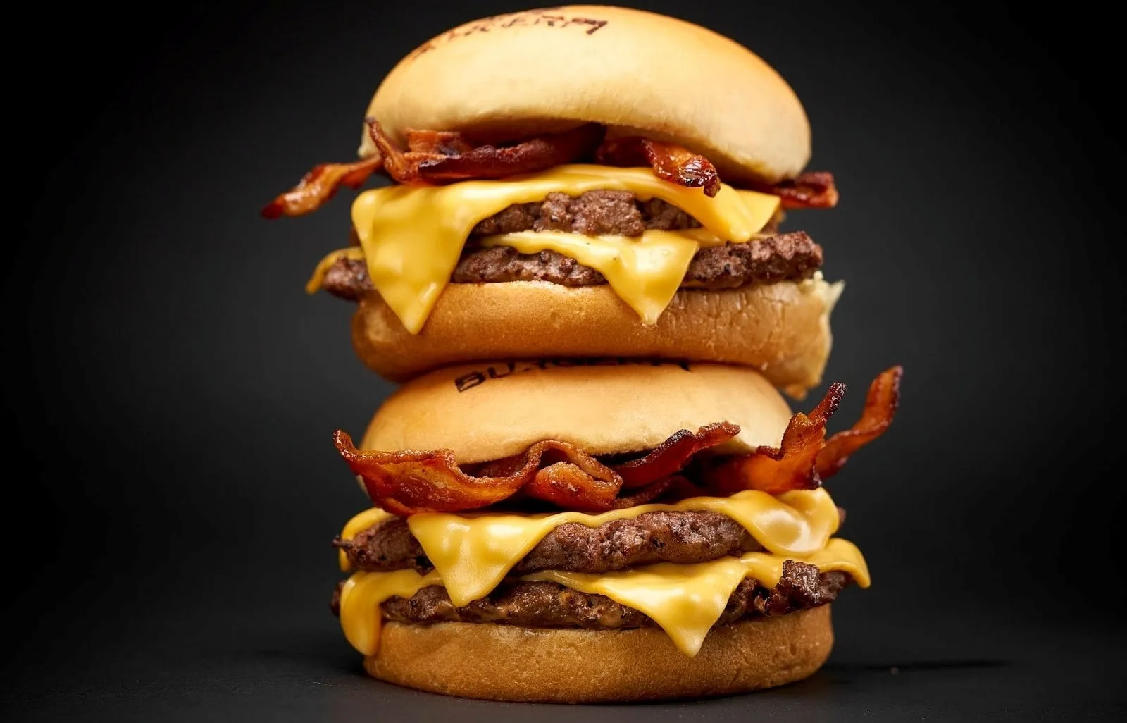 A double bacon cheeseburger with melted cheese and crispy bacon, stacked high on a black background.