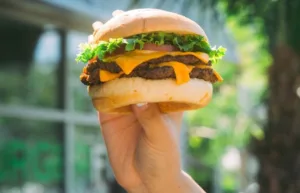 A hand holding a double hamburger with lettuce, tomato, and melted cheese in an outdoor setting.