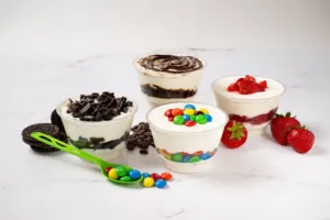 A delightful assortment of creamy desserts topped with Oreos, chocolate drizzle, M&M’s, and fresh strawberries, served on a white marble background.