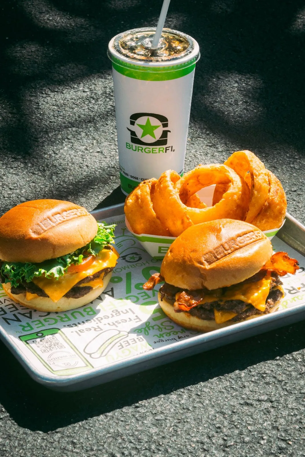 A tray with two delicious burgers, crispy onion rings, and a refreshing soft drink, served outdoors under sunlight.