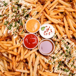 A mountain of crispy french fries with a variety of dipping sauces, including ketchup, mustard, and a creamy garlic aioli.