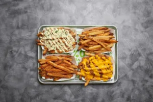 Four different styles of French fries are presented in individual paper containers on a tray. Each serving features unique toppings and sauces.