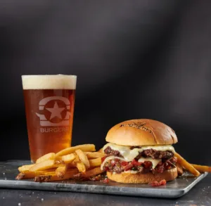 A juicy burger patty topped with melted cheese, served with fries and a pint of beer on a dark background.