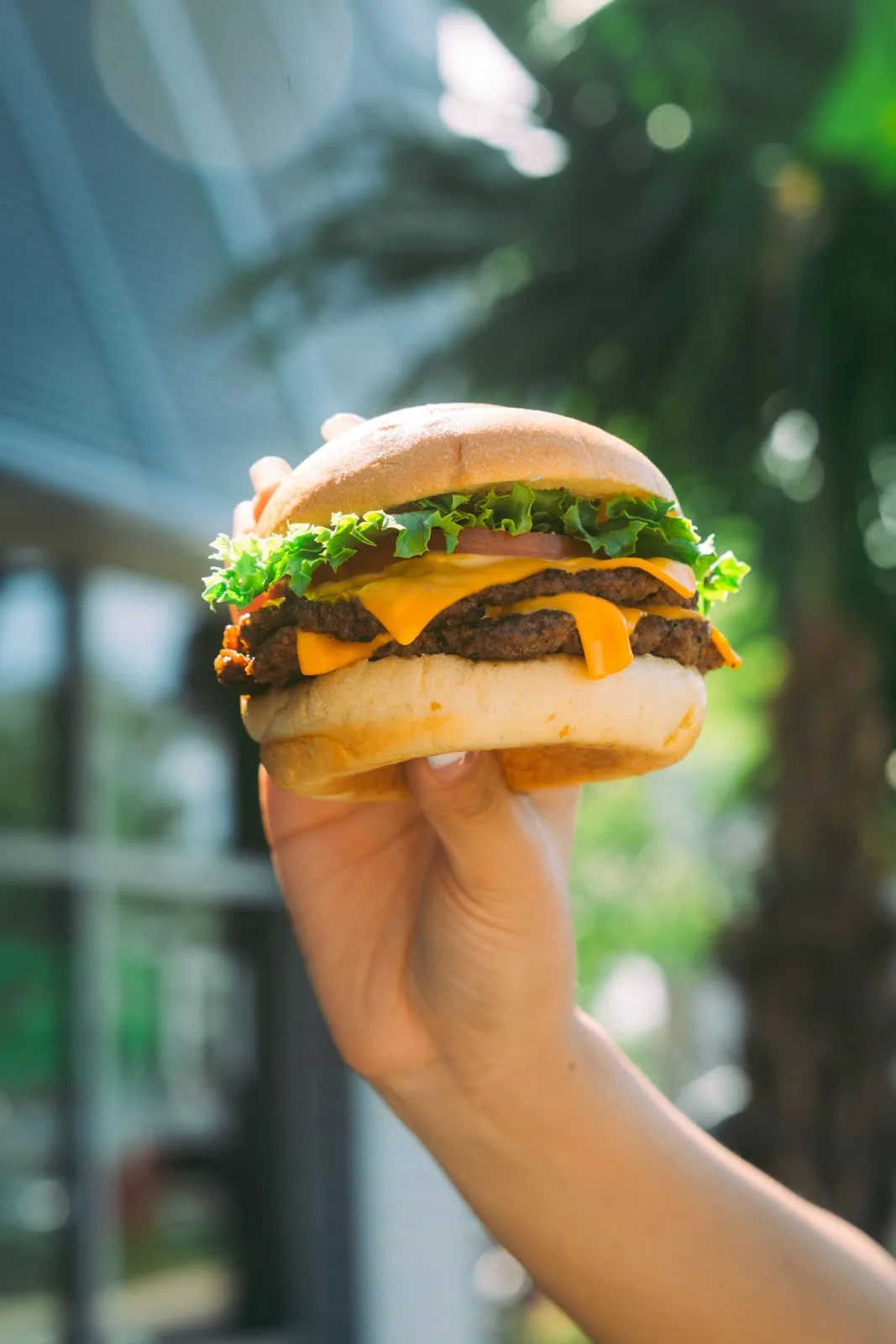 A juicy double cheeseburger with two melted cheese slices, crisp lettuce, and a tomato, held in a hand against a sunny outdoor background.