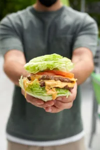 A close-up of a juicy cheeseburger with two burger patties, melted cheese, lettuce, and tomato, held in a hand against a blurred outdoor background.