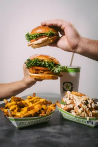 Two people holding burgers, with two baskets of french fries on a table.