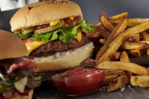 A cheeseburger with cheese, lettuce, tomato, onion, pickles, and mustard on a bun, served with a side of french fries and a small dish of ketchup.