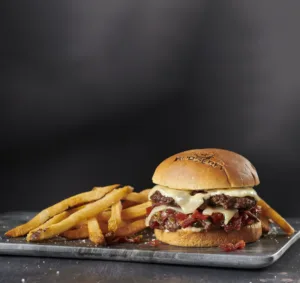 A double cheeseburger with two beef patties, cheese, peppers, onions, and lettuce on a bun, served with a side of french fries.