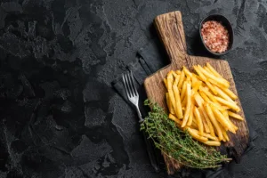 A plate of crispy golden French fries on a wooden board, garnished with fresh thyme and sprinkled with coarse salt.